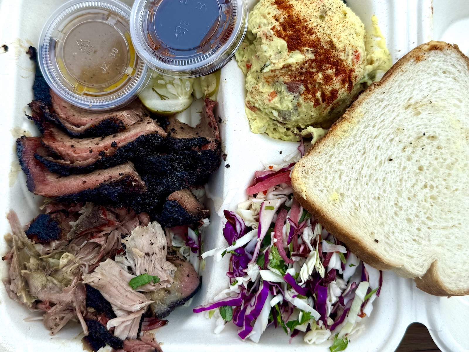 A takeout container of sliced brisket, pulled pork, sauce containers, potato salad, coleslaw, and a slice of white bread