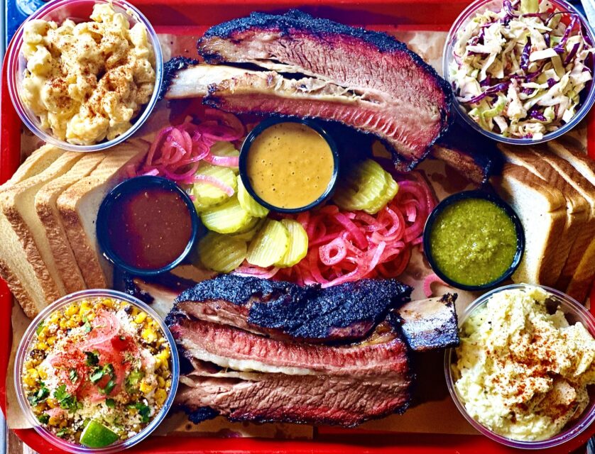 A barbecue platter with two smoked beef ribs, pickled vegetables, cups with side dishes, and stacks of sliced white bread