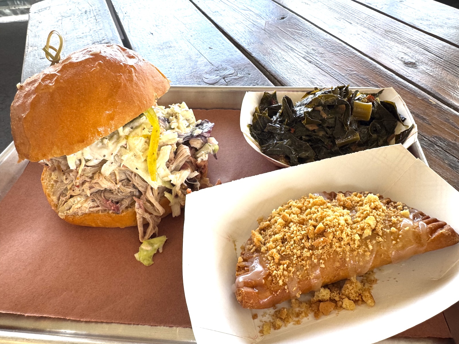 A pulled pork and coleslaw sandwich with a peanut-topped hand pie and collard greens in paper boats on a metal tray with a paper liner.
