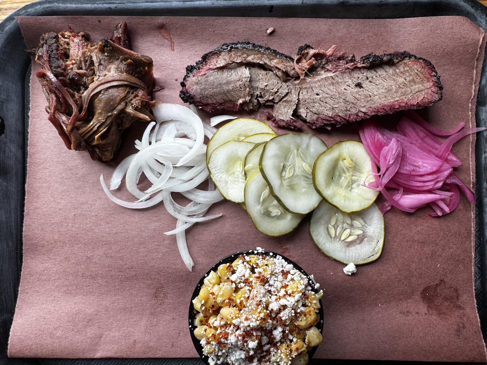 Shredded smoked meat, slices of brisket, pickled white and red onions, pickle slices, and Mexican-style corn in a ramekin on a tray with a paper liner.
