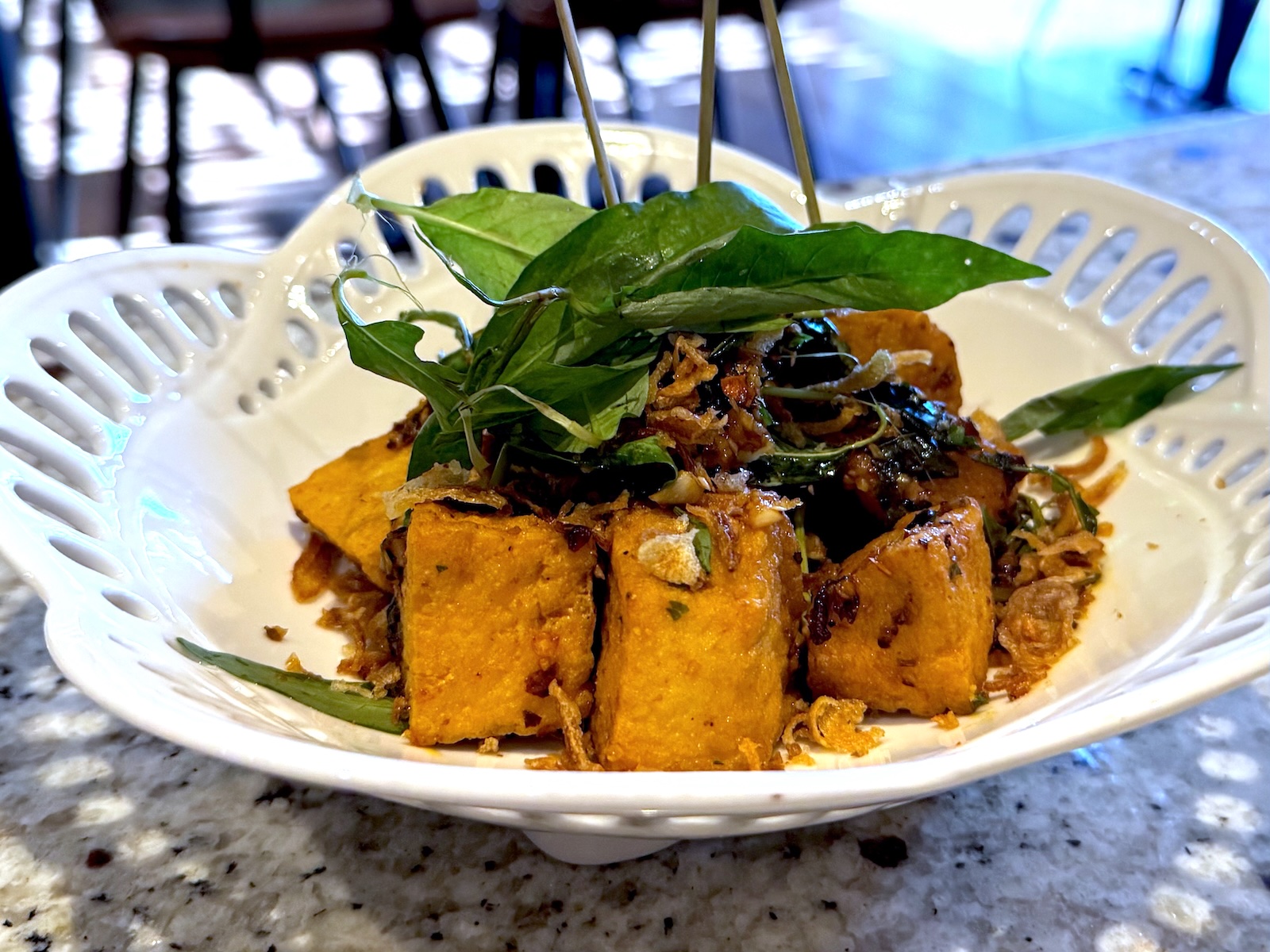 A white bowl with scalloped edges holding cubes of sauce-covered tofu on skewers with fresh herbs
