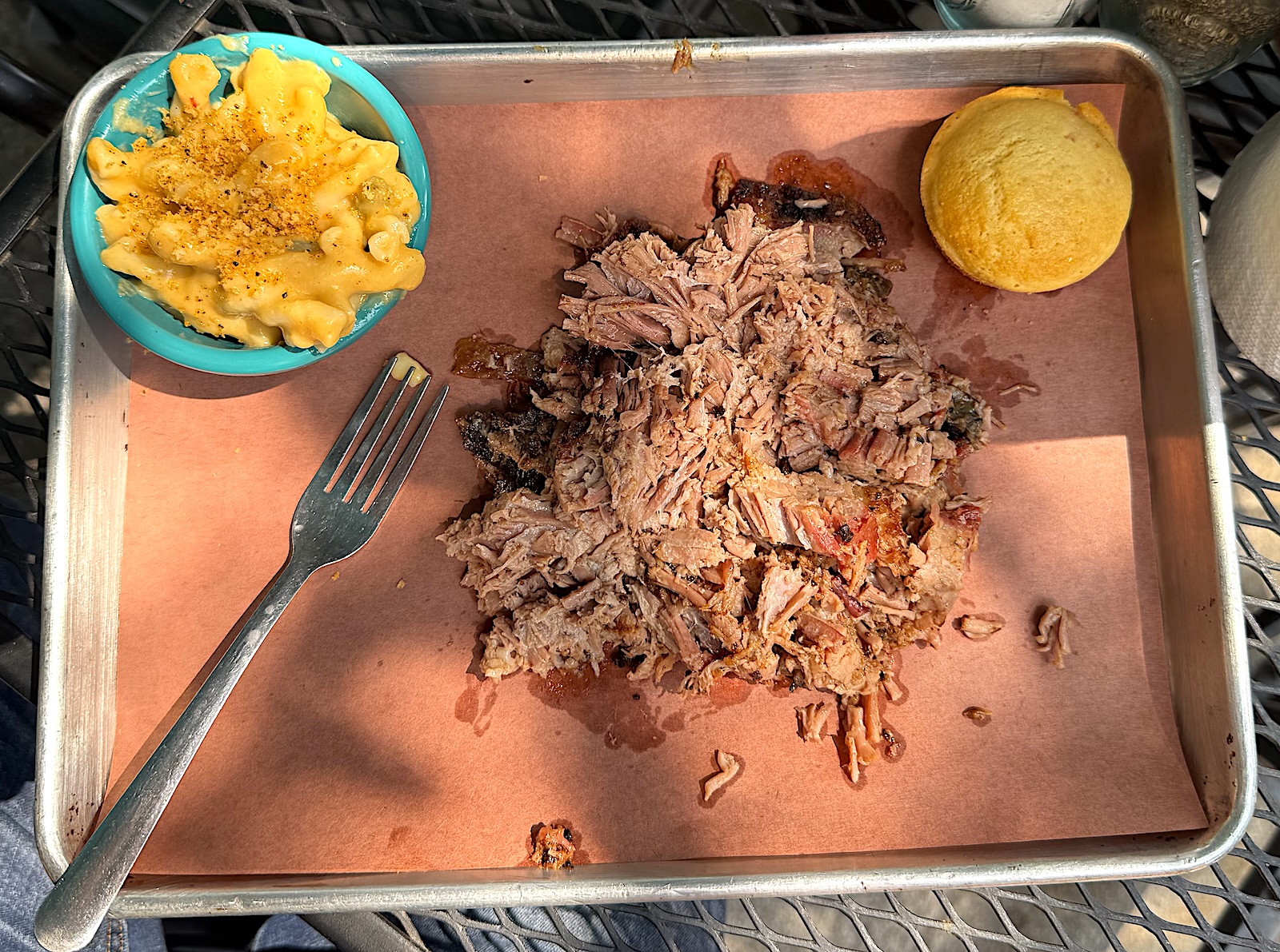 A pile of shredded pork, a cornbread muffin, a ramekin of macaroni and cheese, and a fork on a paper-lined metal tray.