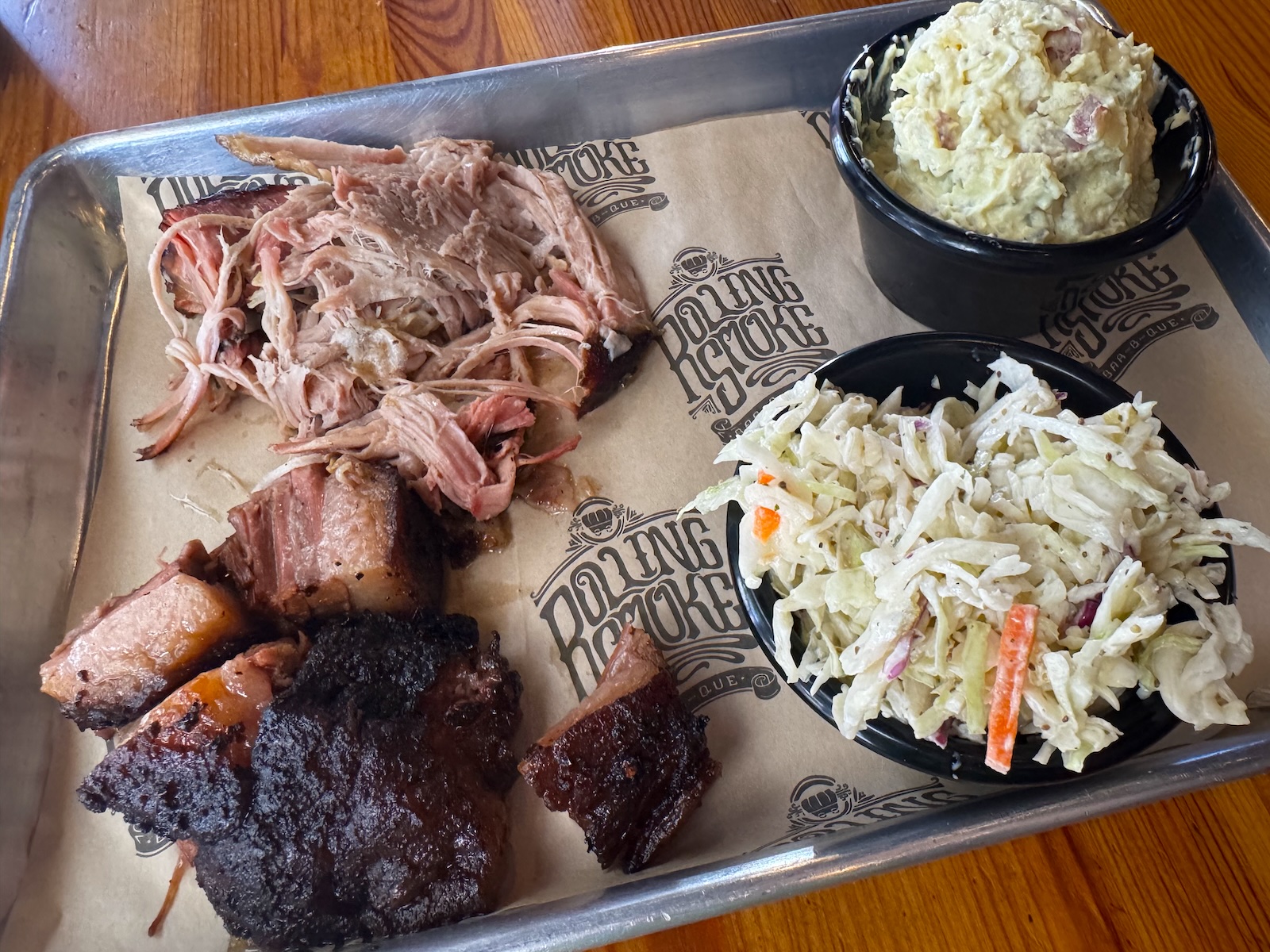 Smoked pork shoulder, pieces of smoked brisket, and ramekins of coleslaw and potato salad on a metal tray with a paper liner labeled Rolling Smoke Barbecue.