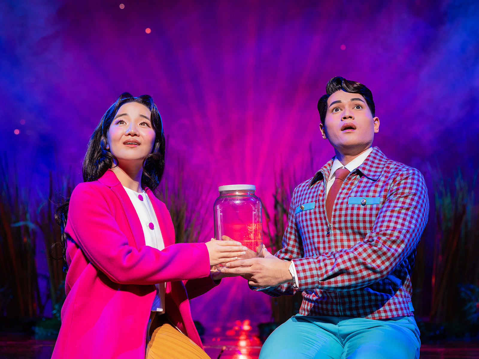 A woman in a pink jacket and a man in blue pants hold a jar in a production of “Maybe Happy Ending”