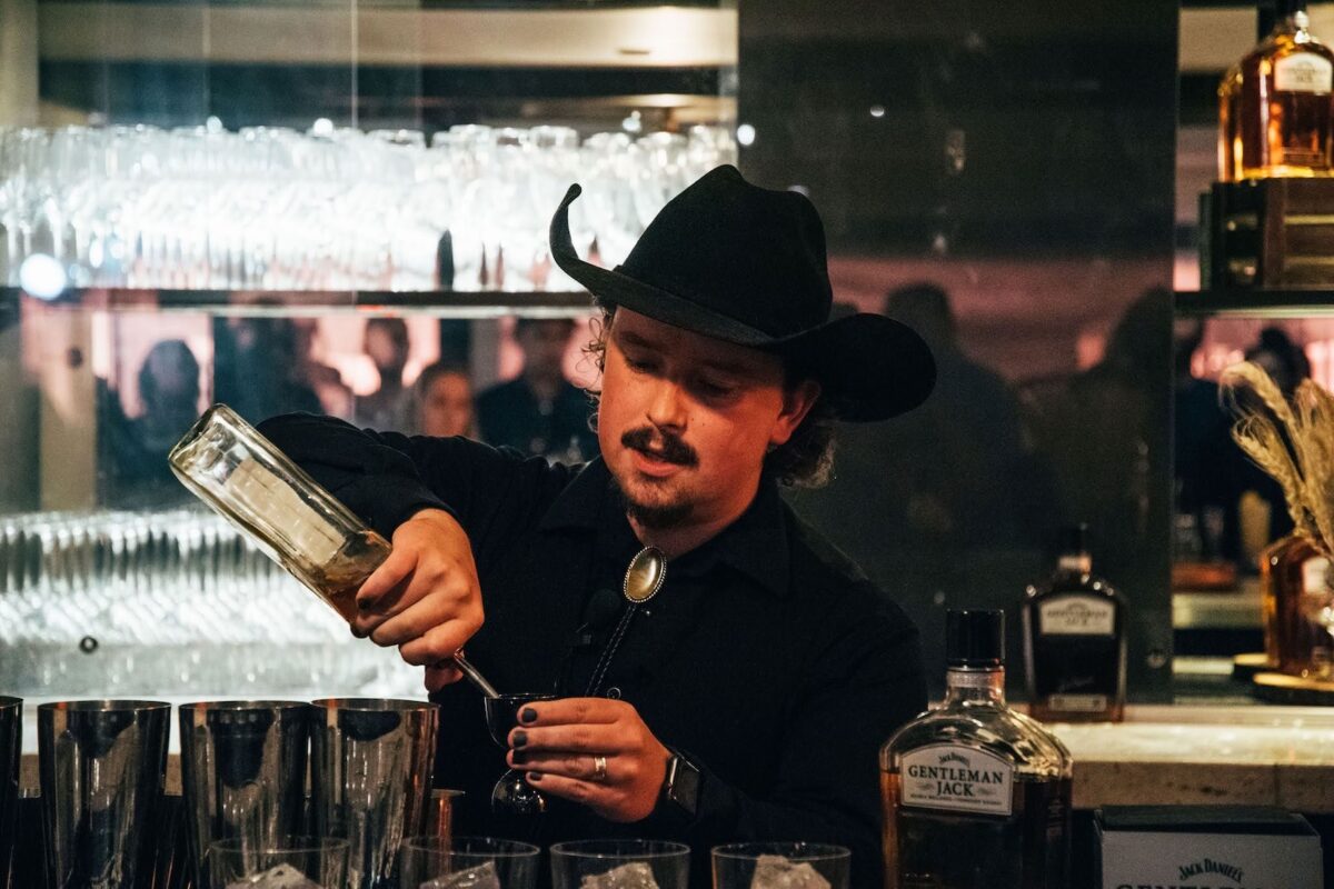 A bartender in a black cowboy hat pours liquor from a bottle.