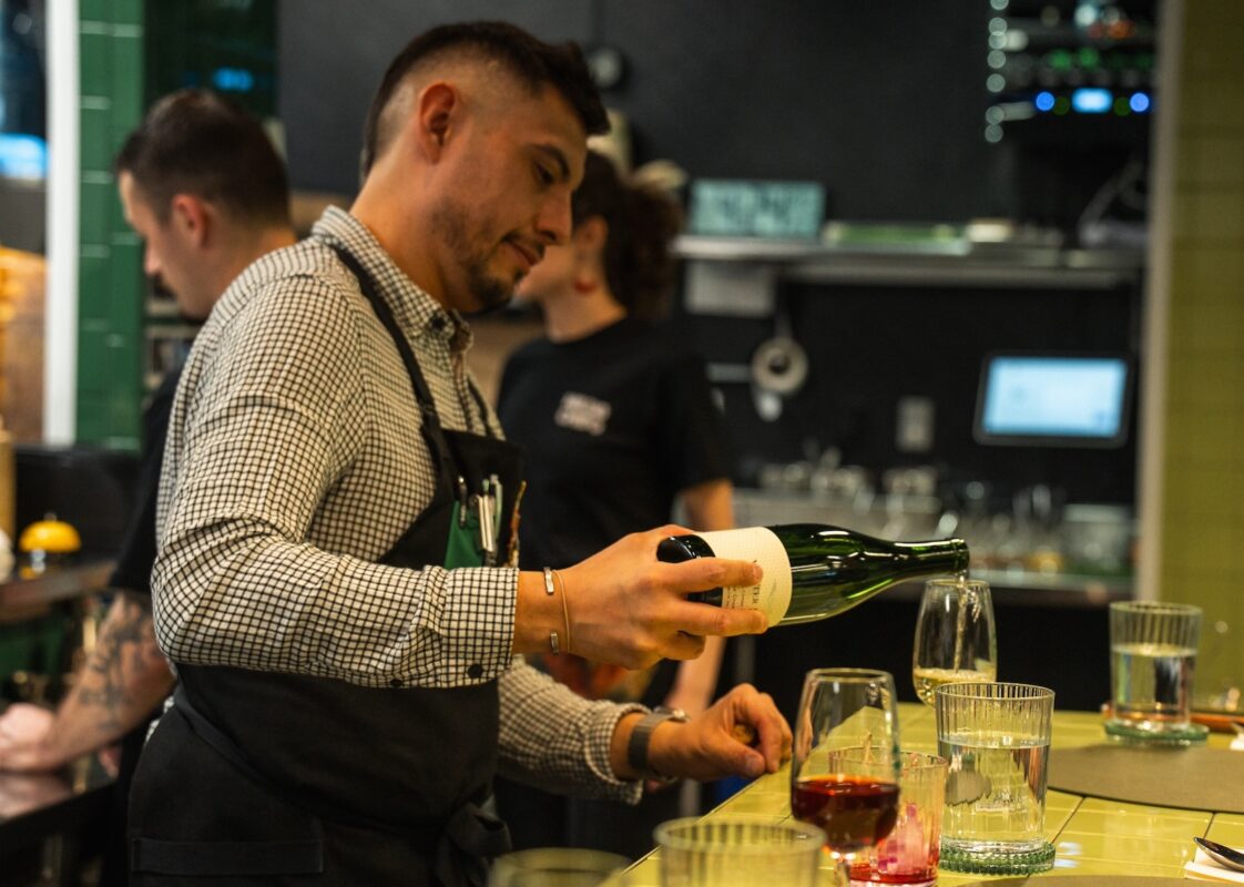 A bartender wearing an apron pours a glass of red wine.