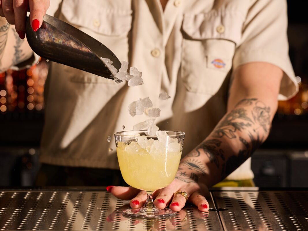A bartender with tattooed arms and a tan shirt adds ice to a cocktail goblet using a metal scoop
