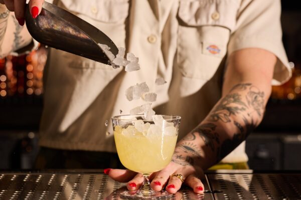 A bartender with tattooed arms and a tan shirt adds ice to a cocktail goblet using a metal scoop